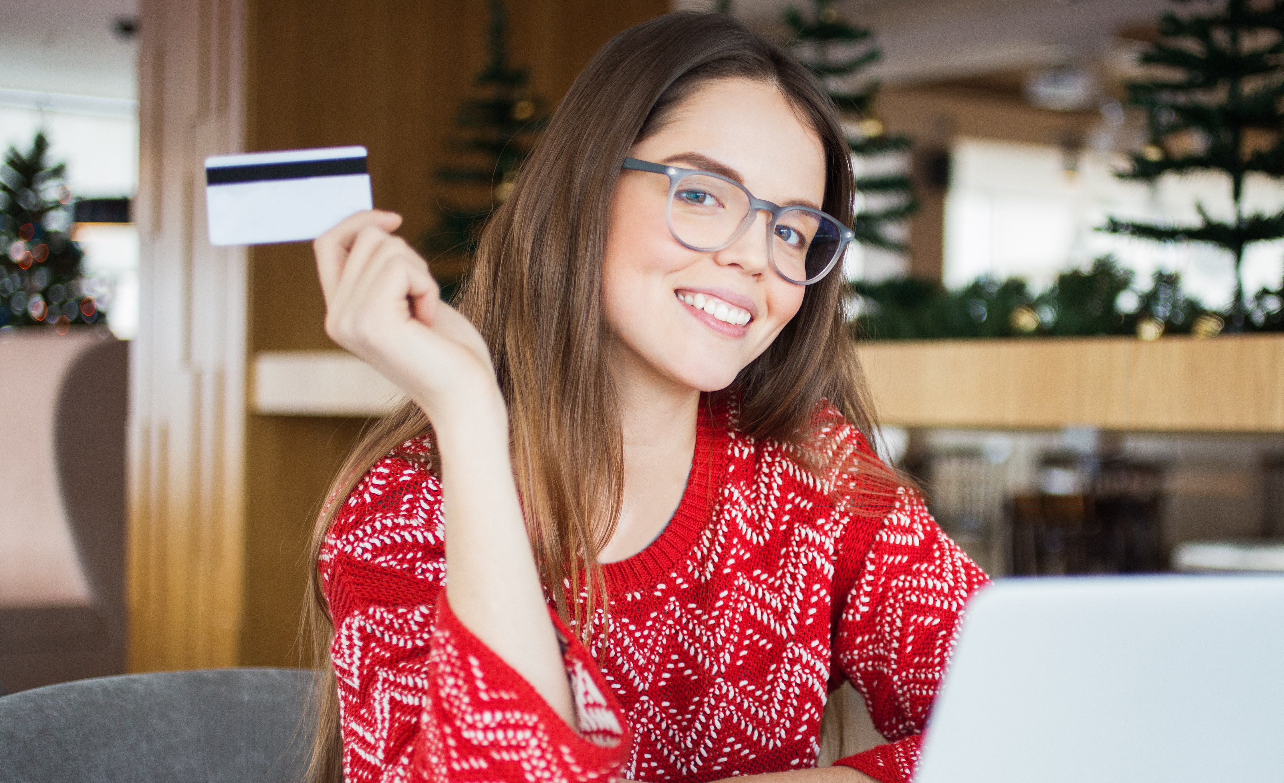 Portrait of positive young Caucasian woman wearing glasses and red knit sweater sitting at laptop in cafe or at home holding credit card for online shopping, looking at camera and smiling